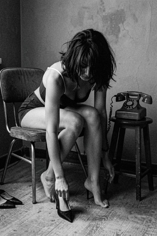 In a moody room, she sits on a retro chair while bending towards the
